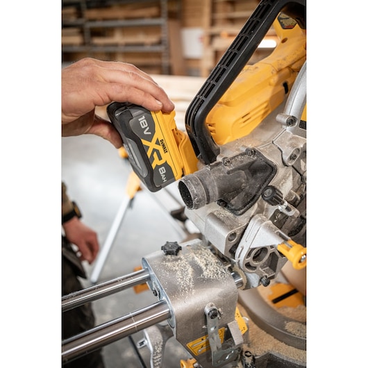 Close-up of a person inserting an 18V XR DEWALT battery into a DEWALT power tool, in a workshop setting with visible sawdust.
