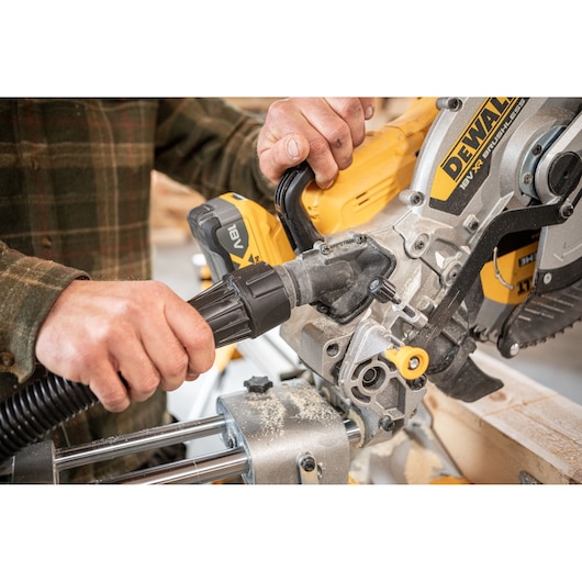Close-up of a person connecting a vacuum hose to a DEWALT 18V cordless miter saw, with sawdust visible on the tool and a piece of wood being cut.
