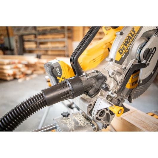 Close-up view of a DEWALT DCS782N cordless mitre saw in a woodworking workshop, highlighting the dust extraction hose attached to the saw.