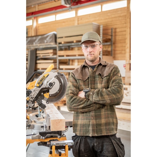 A person with a blurred face stands with arms crossed next to a DEWALT DCS782N miter saw in a woodworking shop. The saw is positioned on a stand and cutting a wooden beam. The background shows shelves and wooden materials.