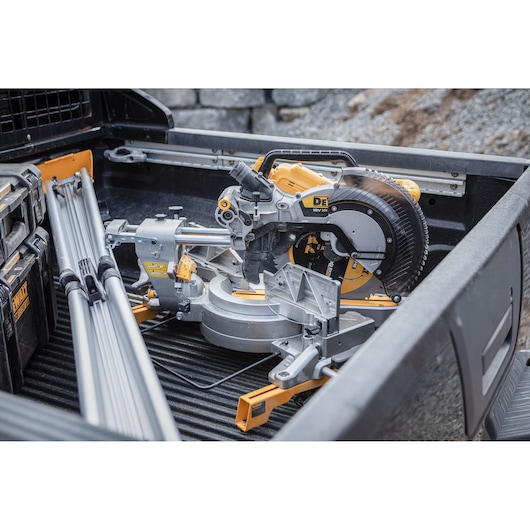 A DEWALT miter saw with a yellow and black design is placed in the bed of a pickup truck along with metal rods and DEWALT tool cases. The saw appears ready for transport to a worksite.