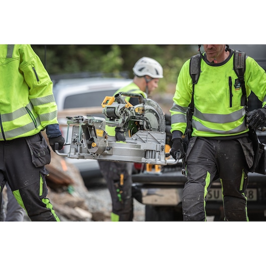 Two construction workers in bright safety clothing are carrying a DEWALT DCS782N miter saw at a work site. The workers’ faces are blurred for privacy. The saw is silver and yellow, and the background shows vehicles and building materials.