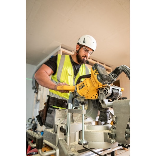 A person wearing a safety vest and helmet operating a DEWALT DCS782N power saw in a construction setting. The DEWALT branding and battery pack are visible.