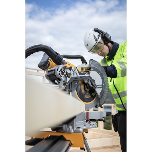A DEWALT DCS782N cordless mitre saw is being used outdoors by a person wearing safety gear and high-visibility clothing. The saw is cutting through a piece of wood on a construction site.
