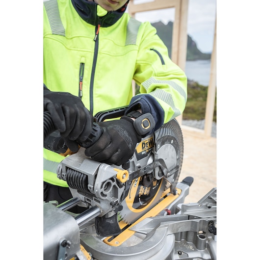 A person wearing a high visibility jacket and gloves is operating a DEWALT DCS782N mitre saw at a construction site with a scenic outdoor background.