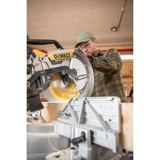 A person operating a DEWALT 18V XR brushless mitre saw, cutting a piece of wood in a workshop. The DEWALT saw is prominent in the foreground with the saw blade spinning, and the operator's face is not clearly visible.