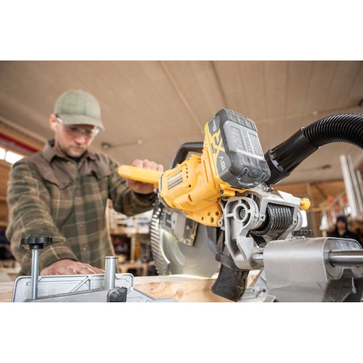 Close-up of a DEWALT DCS782N mitre saw being operated by a person in a workshop. The person is wearing a plaid shirt and cap. The focus is on the saw cutting a piece of wood, with the battery and dust extraction hose visible.
