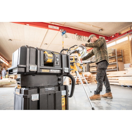 A DEWALT brushless 54V power tool and storage system is in the foreground in a workshop. In the background, a person is operating a DEWALT miter saw, surrounded by wooden planks and workshop equipment.