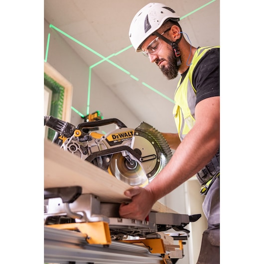 A person wearing safety equipment operates a DEWALT DCS782N mitre saw to cut a wooden plank, with a green laser guide visible in the background.