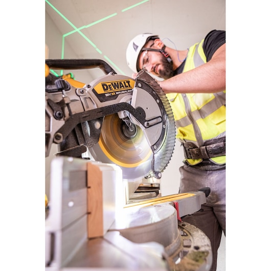 A person wearing safety equipment operates a DEWALT 18V XR brushless miter saw to cut a piece of wood indoors.