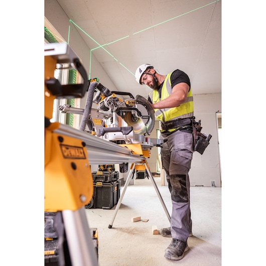 A worker wearing a safety helmet and high-visibility vest uses a DEWALT miter saw (SKU: DCS782N) to cut wood in an indoor construction site. The saw is mounted on a sturdy yellow stand, and laser lines are visible on the ceiling for precise measurement. Various DEWALT toolboxes and accessories are scattered in the background.