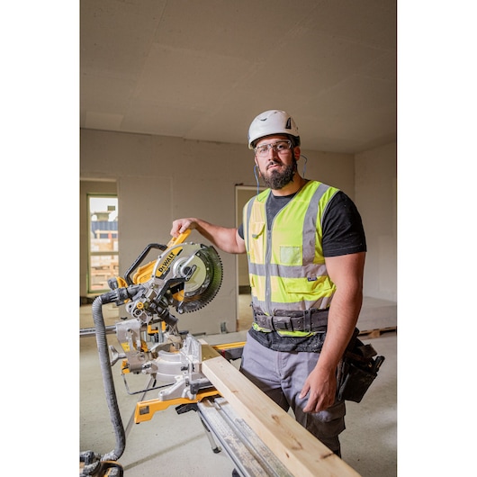 A construction worker wearing a safety helmet and high-visibility vest operates a DEWALT DCS782N mitre saw, cutting a wooden plank inside a building under construction.