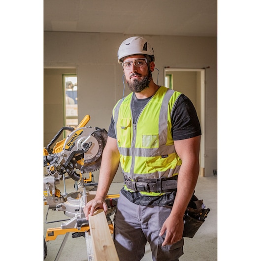 A construction worker wearing a safety helmet and high-visibility vest stands next to a DEWALT DCS782N miter saw. The worker is holding a wooden plank, and the saw is set up indoors, suggesting use in a building or renovation environment.