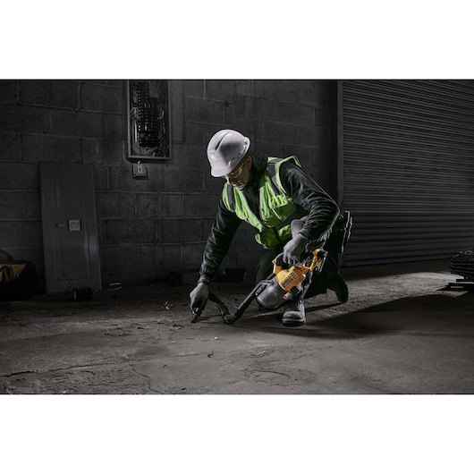 A construction worker wearing a high-visibility vest and hard hat uses a DEWALT portable vacuum cleaner (model DCV501LN) to clean debris from the floor of an industrial site.