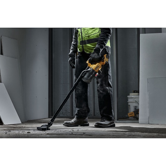 A person wearing work gloves and a safety vest is using a yellow and black DEWALT cordless handheld vacuum cleaner to clean construction debris from a floor at a building site.