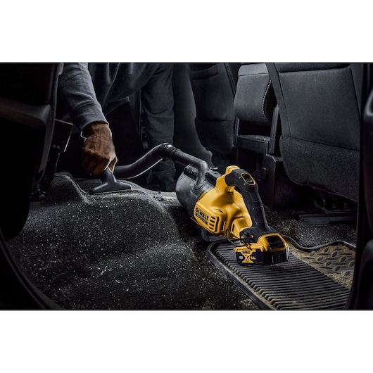 A person using a DEWALT cordless handheld vacuum cleaner to clean the floor of a car's interior.