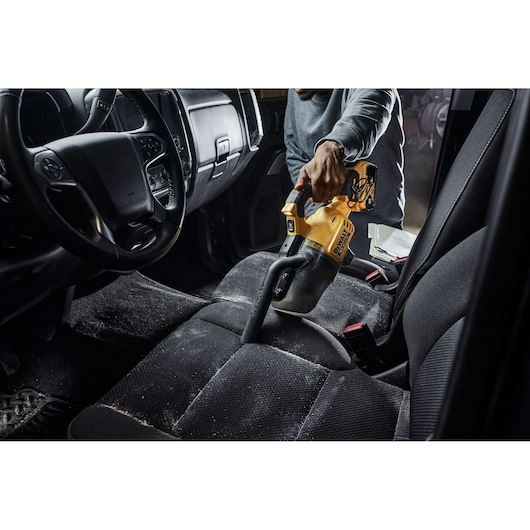 A person is using a DEWALT handheld vacuum cleaner to clean the front seat of a car, removing dust and debris from the upholstery.