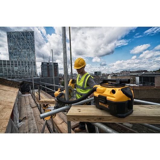 A DEWALT DCV584L vacuum cleaner is placed on a wooden platform at a construction site, with a worker in a yellow hard hat and high-visibility vest operating a tool. Tall city buildings and a partly cloudy sky are visible in the background.