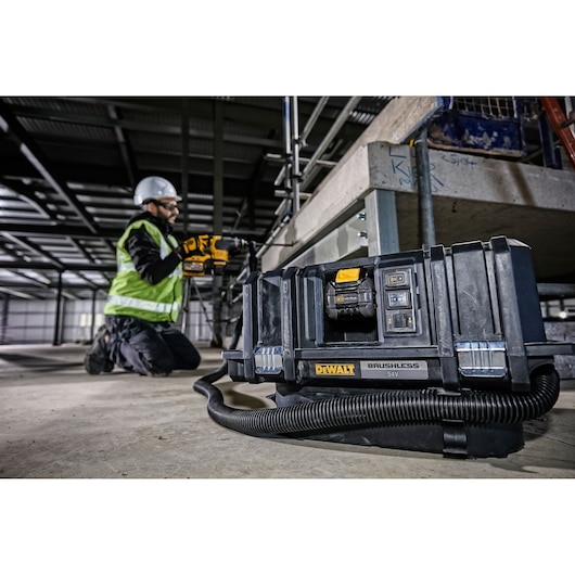 A DEWALT DCV586MT2 brushless 54V vacuum cleaner is positioned on the floor of a construction site, with its hose coiled in front. In the background, a worker wearing a safety vest and helmet is kneeling, operating a power tool near scaffolding. The scene takes place in an industrial indoor setting, highlighting the use of DEWALT equipment for professional applications.