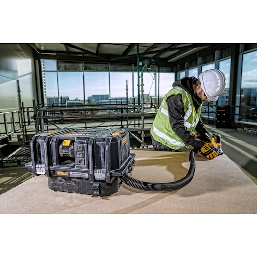 A DEWALT brushless portable dust extractor (model DCV586MT2) is placed on a worktable in a construction site. A construction worker wearing a white hard hat, safety vest, and gloves is using a power tool that is connected to the dust extractor with a hose.