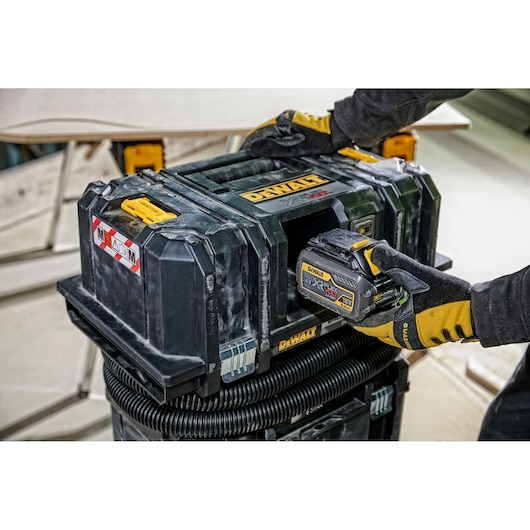 A person wearing yellow and black gloves is handling a DEWALT cordless dust extractor. The device is rectangular with a rugged black body, and it is equipped with an 18V XR FLEXVOLT battery pack. The vacuum is placed on top of a hose and is shown in a construction or workshop setting.
