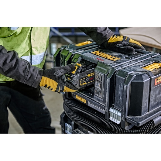 A person wearing yellow and black gloves is inserting a DEWALT 18V XR FLEXVOLT battery into a black DEWALT brushless vacuum unit at a worksite.