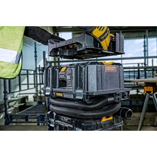 A person wearing work gloves and a safety vest is handling a DEWALT portable dust extractor vacuum in an industrial or construction setting. The vacuum has a black and yellow design, with a coiled hose around the base and modular storage compartments.