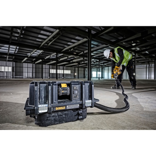 A DEWALT brushless vacuum cleaner is shown on the floor of a large industrial building under construction. It is connected by a hose to a worker operating a power tool in the background.