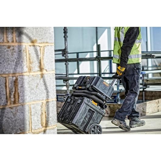 A construction worker wearing a high-visibility vest and gloves is pulling a DEWALT mobile toolbox and vacuum system with wheels at a building site. The worker is only partly visible, and the scene is set outdoors with scaffolding and a brick wall in the background.