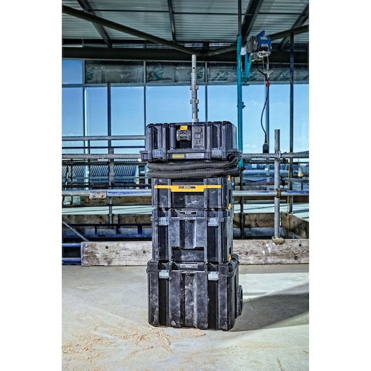 DEWALT DCV586MT2 portable stack of black tool storage boxes with a dust extractor unit, shown on a construction site with pipes and scaffolding in the background.