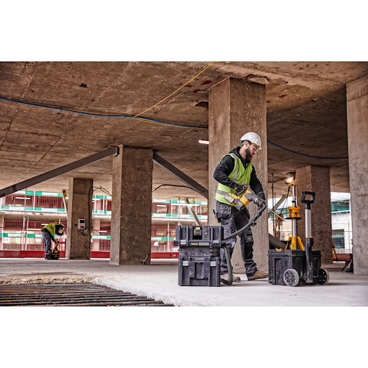 A worker wearing a high visibility vest and a hard hat uses a DEWALT DCV586MT2 dust extractor with attached hose on a construction site. The extractor is surrounded by DEWALT tool storage boxes, and another worker can be seen in the background. The environment features concrete pillars and beams.