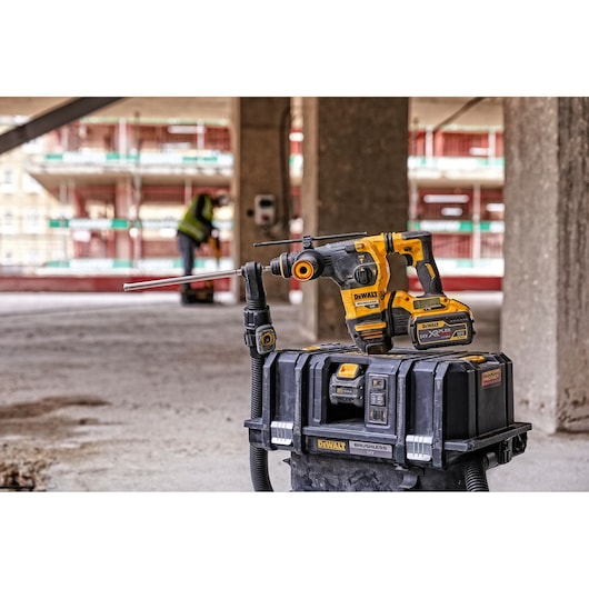 A DEWALT cordless rotary hammer drill with attached dust extraction system is placed on top of a DEWALT storage box on a construction site. In the background, a person in a safety vest is working.
