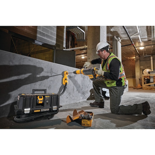 A construction worker wearing safety gear uses a DEWALT cordless dust extractor system and rotary hammer drill to bore into a concrete wall in an industrial setting. The DEWALT tool case and accessories are placed on the ground nearby.