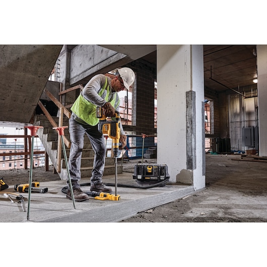 A worker in a construction site wearing a safety vest and helmet is using a DEWALT cordless power tool to drill into a concrete surface. The DEWALT DCV586MT2 dust extractor is visible on the floor beside the worker.