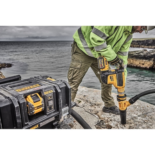 A person wearing a green high-visibility jacket is using a yellow DEWALT cordless rotary hammer drill on a rocky surface near the water, with a DEWALT portable tool box and vacuum system visible in the foreground.