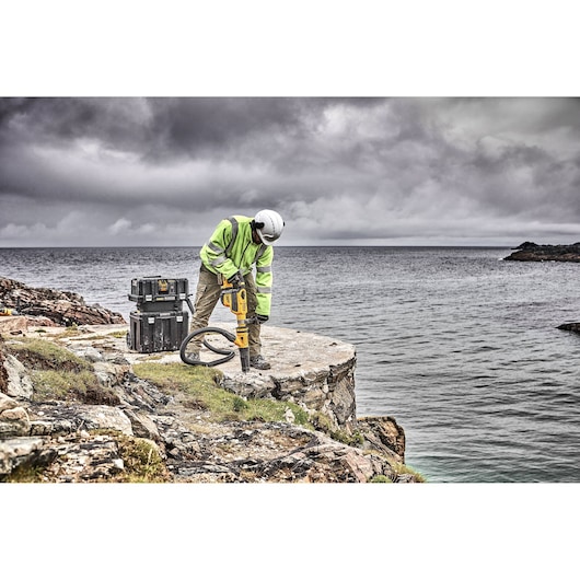 A worker wearing a high-visibility jacket and helmet uses a DEWALT power tool and dust extractor on a rocky coastal cliff, with the ocean and cloudy sky in the background. Two stacked DEWALT toolboxes are next to the worker.