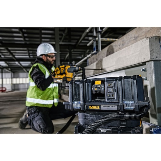 A DEWALT brushless 54V tool with hose attachment is positioned in the foreground on a construction site. In the background, a person wearing a safety helmet and high-visibility vest uses a DEWALT power tool to work on a concrete structure.