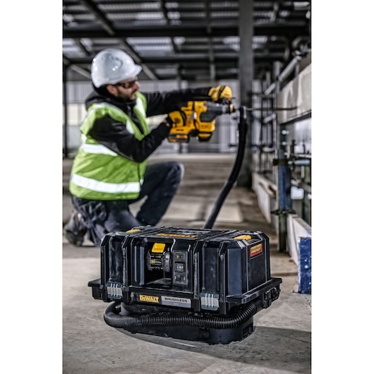 A DEWALT cordless brushless vacuum cleaner (model DCV586MT2) is placed on the floor of a construction site. In the background, a construction worker wearing a high-visibility vest, helmet, and safety gear is kneeling and drilling into a concrete wall. The vacuum is connected via a hose to the worker's tool to help collect dust.