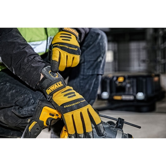 Close-up of a person wearing yellow and black DEWALT gloves, adjusting the wrist strap on one glove while holding a yellow DEWALT power tool. A DEWALT equipment case is visible in the background in an industrial setting.