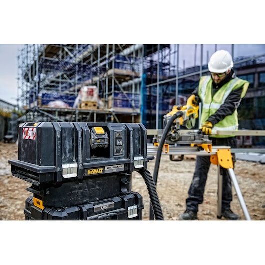 DEWALT cordless dust extractor (model DCV586MT2) in use at a construction site. The tool is connected by a hose to a power saw operated by a worker wearing a high-visibility safety vest, gloves, and a white helmet. Scaffolding and construction materials are visible in the background.