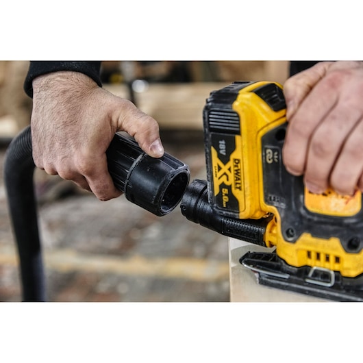Hands connect a hose to a yellow DEWALT DCW200NT power tool for dust extraction during woodworking.