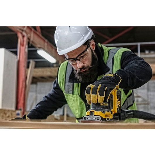 Worker uses DEWALT DCW200N sander on wood, wearing safety gear and gloves at construction site.