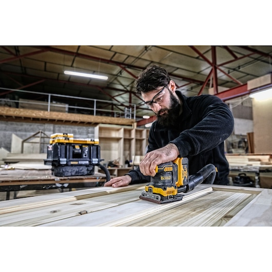 Person using a DEWALT DCW200N sander on wood in a workshop. Tool box visible in background.