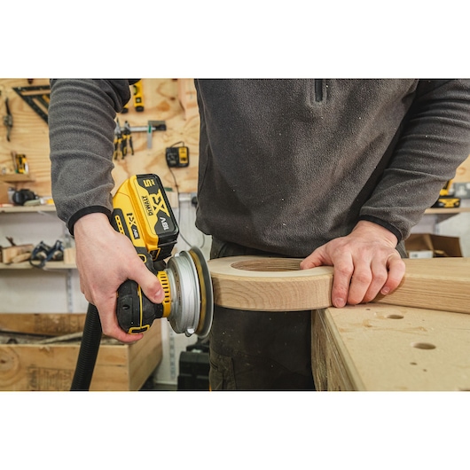 Person uses DEWALT cordless sander to smooth a wooden piece in a workshop.