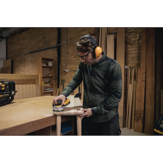 Person using a DEWALT cordless sander on wood in a workshop while wearing ear and eye protection.