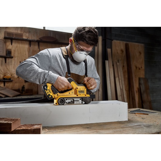 A person using a DEWALT cordless belt sander (SKU: DCW220P2) on a piece of wood in a workshop. The person is wearing safety goggles and an apron, and their face is blurred.