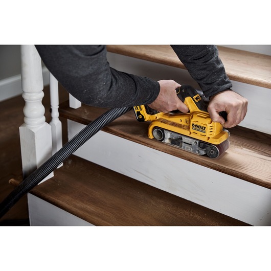 A person is using a DEWALT DCW220P2 yellow cordless belt sander to sand a wooden staircase step. A vacuum hose is attached to the tool for dust extraction.