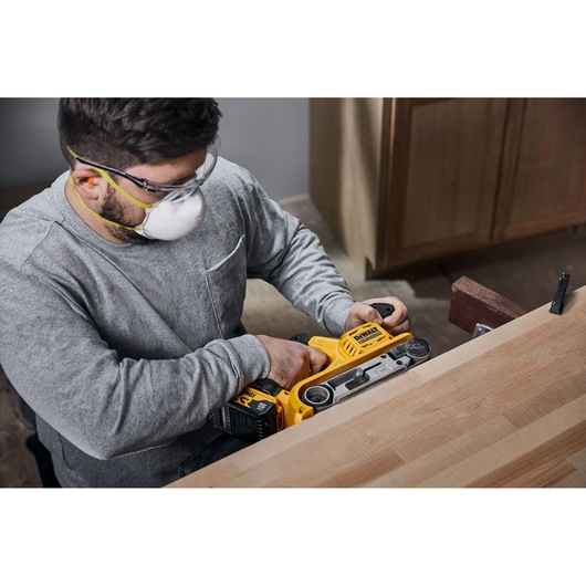 A person using a DEWALT DCW220P2 cordless belt sander to smooth the surface of a wooden table. The user is wearing safety goggles and a dust mask.