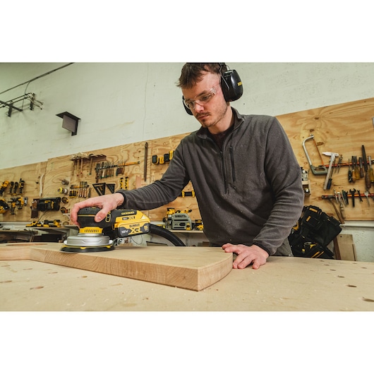 Person sanding wood with a DEWALT DCW230N-XJ sander in a workshop, wearing safety gear.