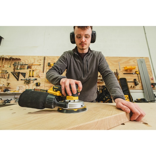 Person using a DEWALT sander on a wooden board in a workshop with tools in the background.